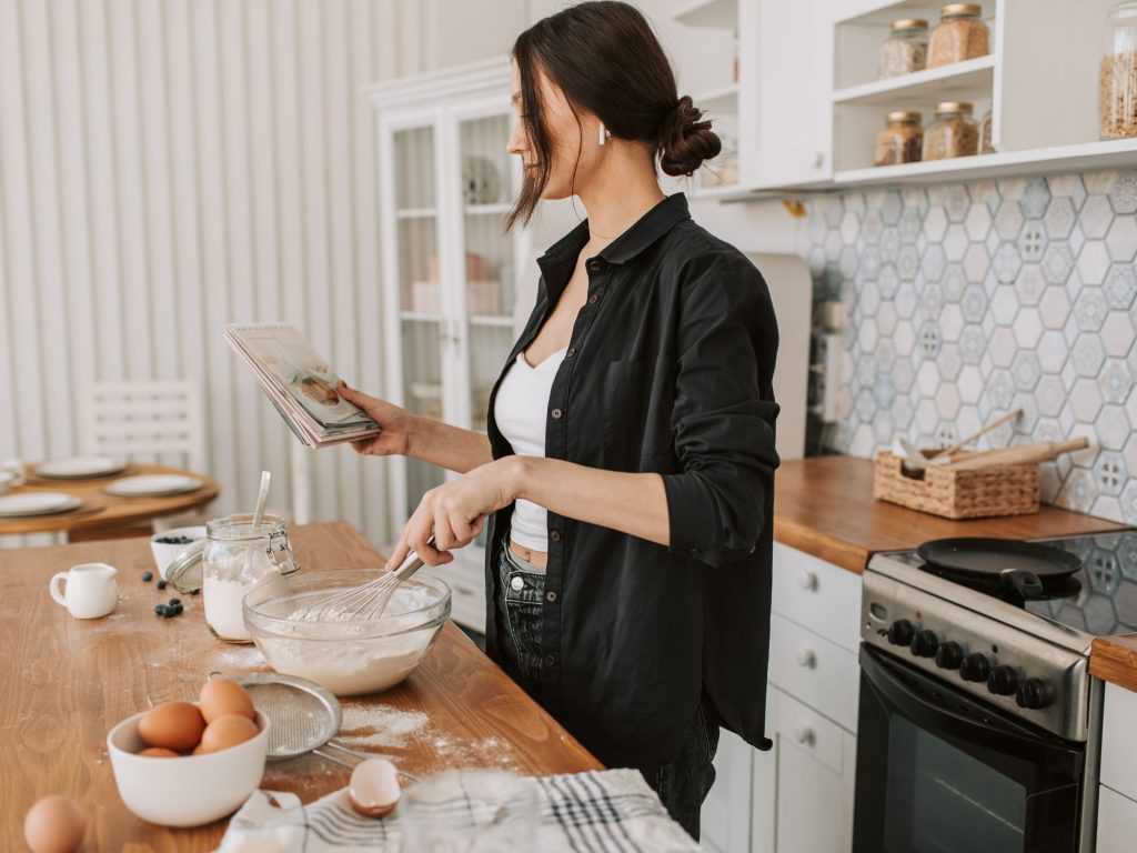 Woman mixing up blueberry pancakes with the ingredients scattered around the counter (Source: Canva).