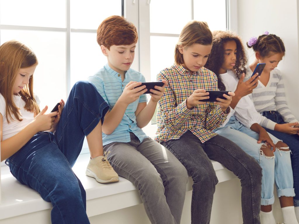 Group of teenagers texting mobile phone messages leaning against urban wall - Row of best student friends using smartphone wearing summer trendy clothing.