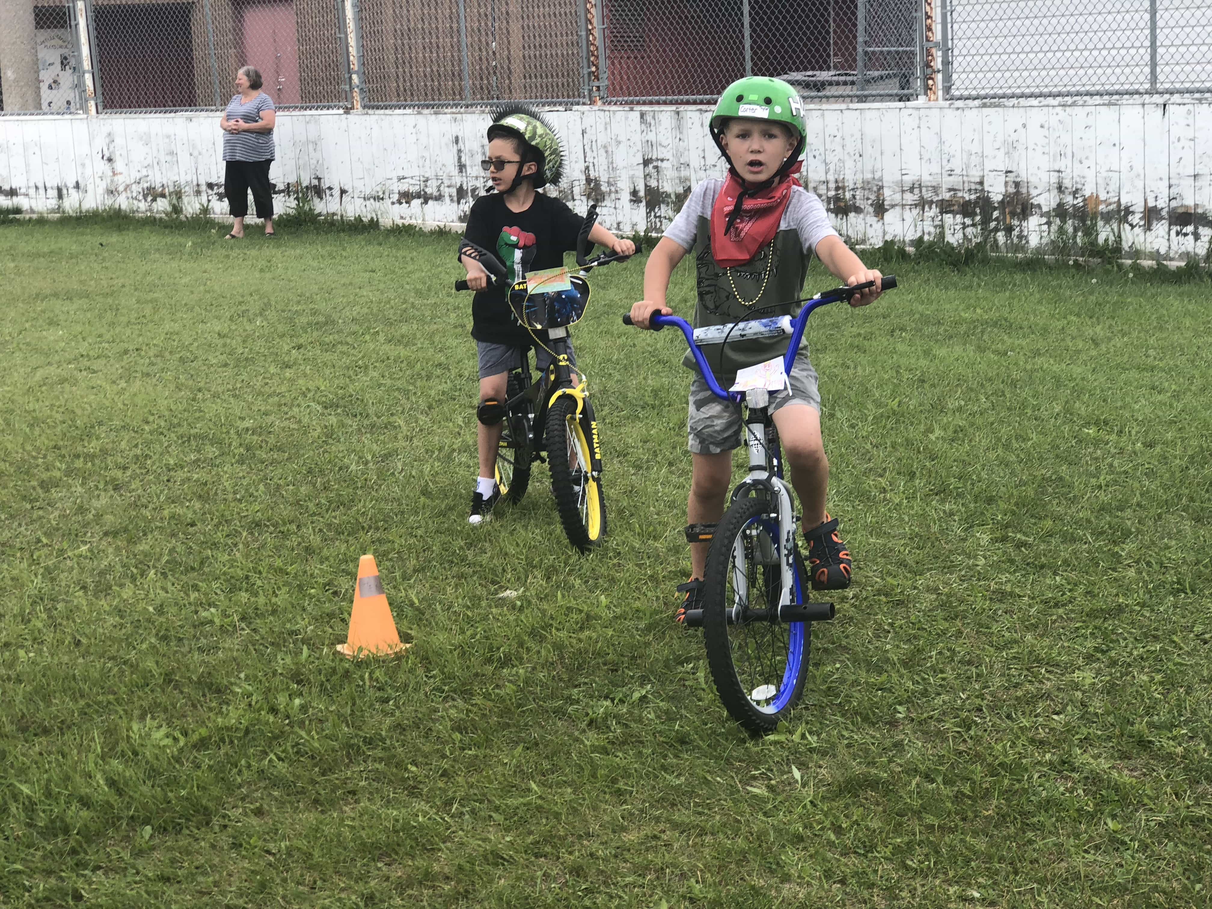 Kids in green helmets riding their bike on grass around a small pylon.