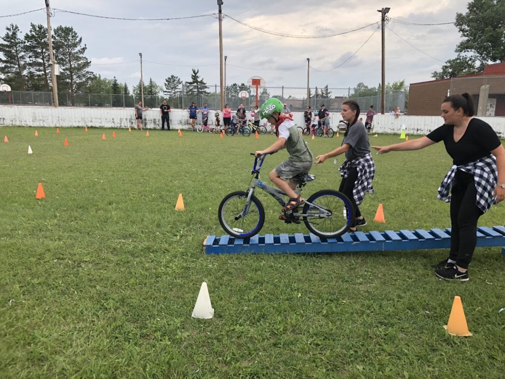 Kid riding his bike across a blue wood bridge in a grass field as part of the Pedalheads summer camp