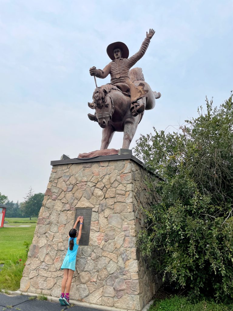 Girl reaches up, giving scale to "The Legacy", the world's largest bucking saddle bronc horse & rider in Ponoka