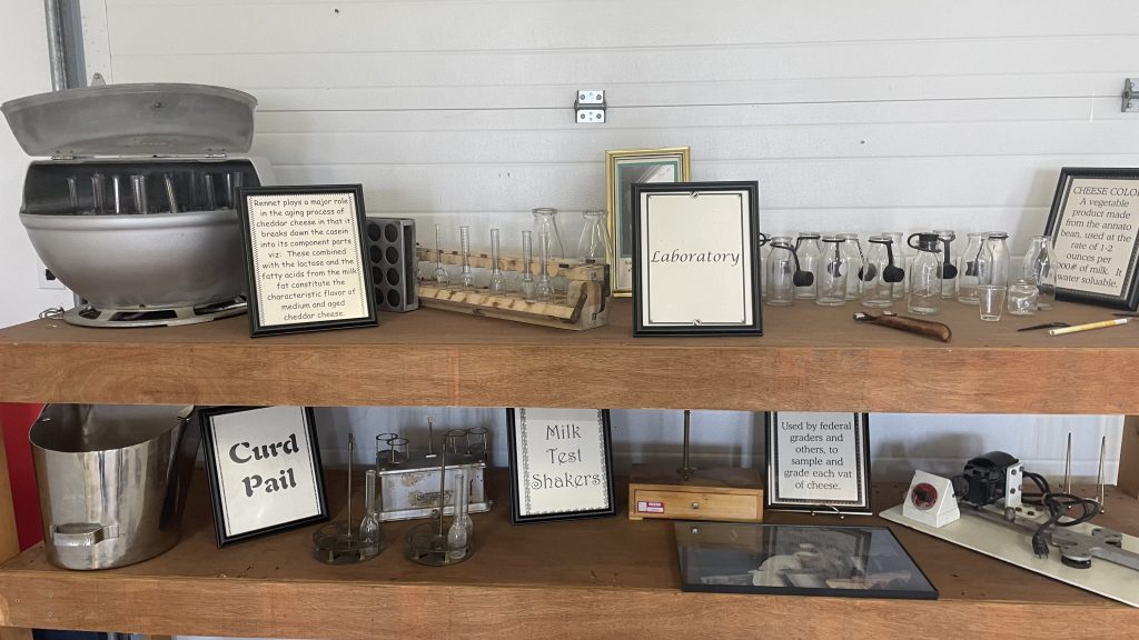 Photo of equipment display at Glenwood Cheese Factory Museum. Framed descriptions read "Renne plays a major role in the agin process of cheddar cheese in that it breaks down the casein into its component parts viz: These combined with the lactose and the fatty acids from the milk fat constitute the characteristic flavour of medium and aged cheddar cheese." And "Cheese Color: A vegetable product made from annatto bean, used at the rate of 1-2 ounces per 1,000# of milk It is water soluble." Also "Laboratory", "Curd Pail", "Milk Test Shakers" and "Used by federal graders and others, to sample and grade each vat of cheese."