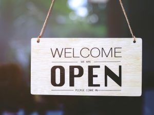 Wood sign reads "Welcome we are Open please come in" over a blurry background.